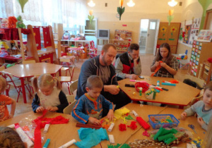 Rodzice i dzieci siedzą przy trzech złączonych stołach, na których leżą kolorowe bibuły.